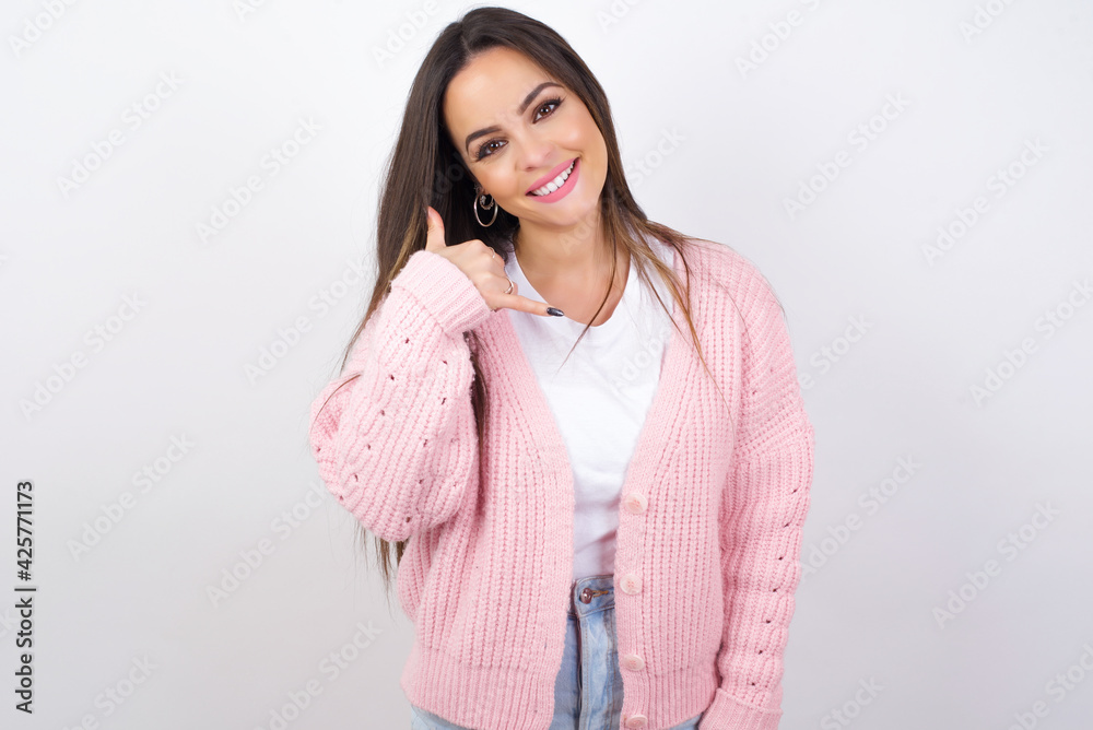 young beautiful caucasian woman wearing pink knitted jacket over white wall smiling doing phone gesture with hand and fingers like talking on the telephone. Communicating concepts. young beautiful caucasian woman wearing pink knitted jacket over white wall smiling doing phone gesture with hand and fingers like talking on the telephone. Communicating concepts.