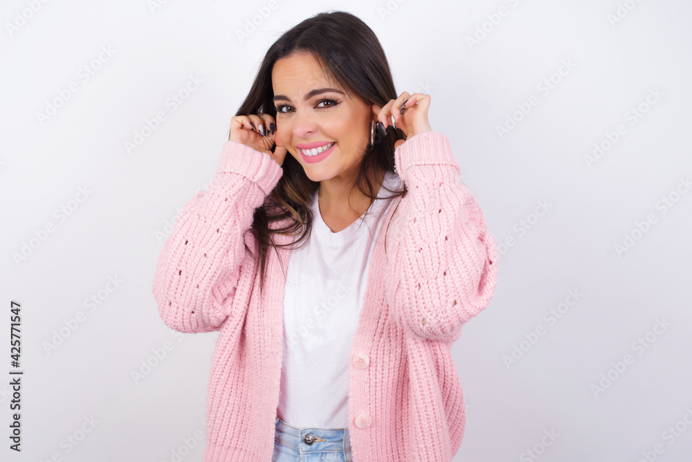 young beautiful caucasian woman wearing pink knitted jacket over white wall covering ears with fingers with annoyed expression for the noise of loud music. Deaf concept. young beautiful caucasian woman wearing pink knitted jacket over white wall covering ears with fingers with annoyed expression for the noise of loud music. Deaf concept.