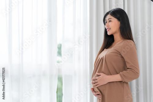 Asian happy pregnant woman is standing beside windows and touching her belly. pregnancy, motherhood, people and expectation concept Free Download Asian happy pregnant woman is standing beside windows and touching her belly. pregnancy, motherhood, people and expectation concept - GFXLoad
