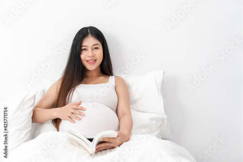 Asian happy pregnant woman is sitting and reading a book on bed and and touching her belly. pregnancy, motherhood, people and expectation concept
