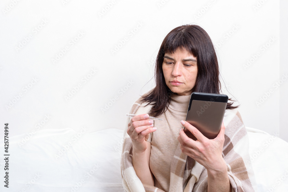 Portrait of a sick woman covered with a blanket holding a thermometer and communicating with the doctor on the tablet. Distance medicine concept. New normal concept. Health concept Portrait of a sick woman covered with a blanket holding a thermometer and communicating with the doctor on the tablet. Distance medicine concept. New normal concept. Health concept