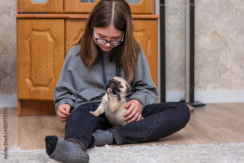 A girl with long hair and glasses sits on the floor and plays with a pug puppy. The puppy frolics and gnaws at the ribbon from the girl's clothes.