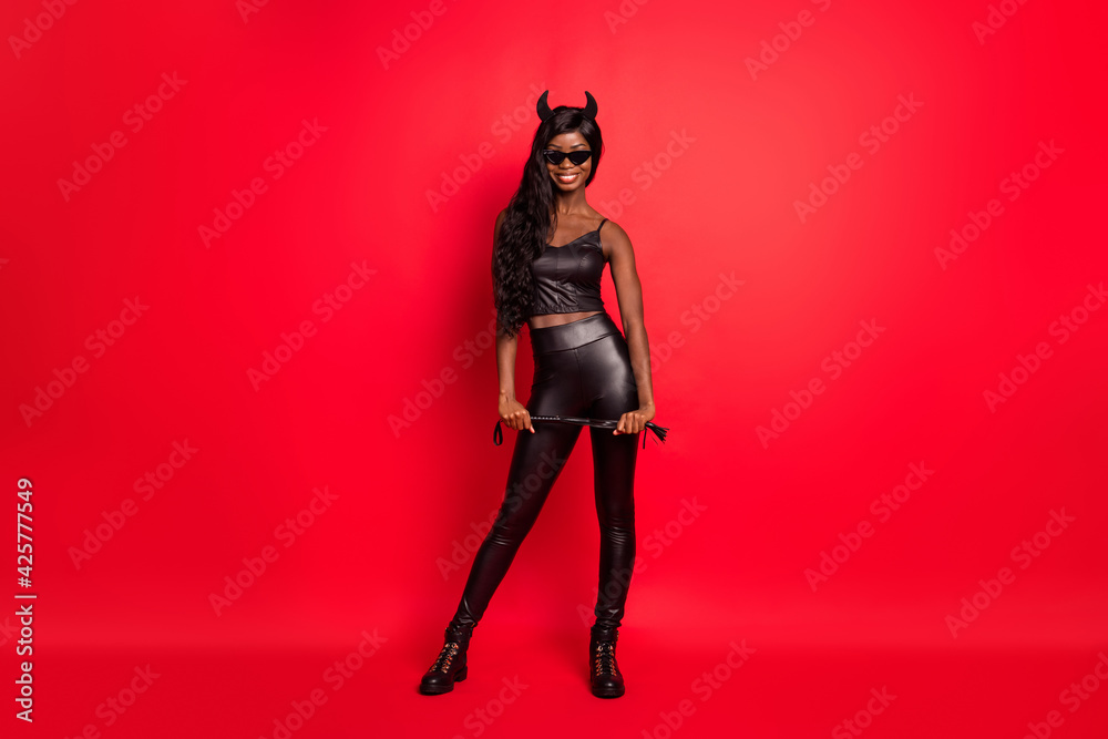 Full length photo portrait of african american horned girl with leather whip isolated on bright red colored background Full length photo portrait of african american horned girl with leather whip isolated on bright red colored background