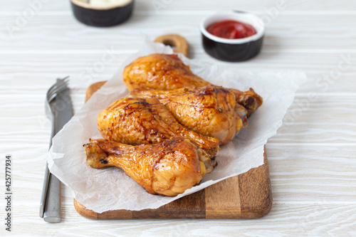 Roasted golden chicken legs drumsticks on baking paper on wooden board served with ketchup and mayonnaise on rustic white wooden table, angle view 