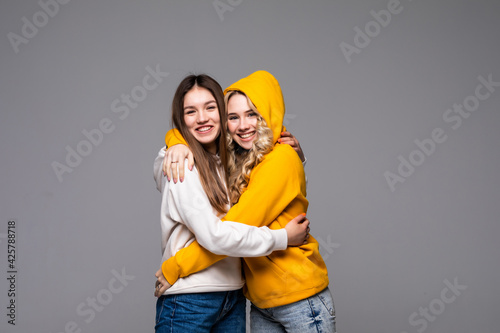 Two beautiful happy smiling young women hugging as best friends, isolated on gray background