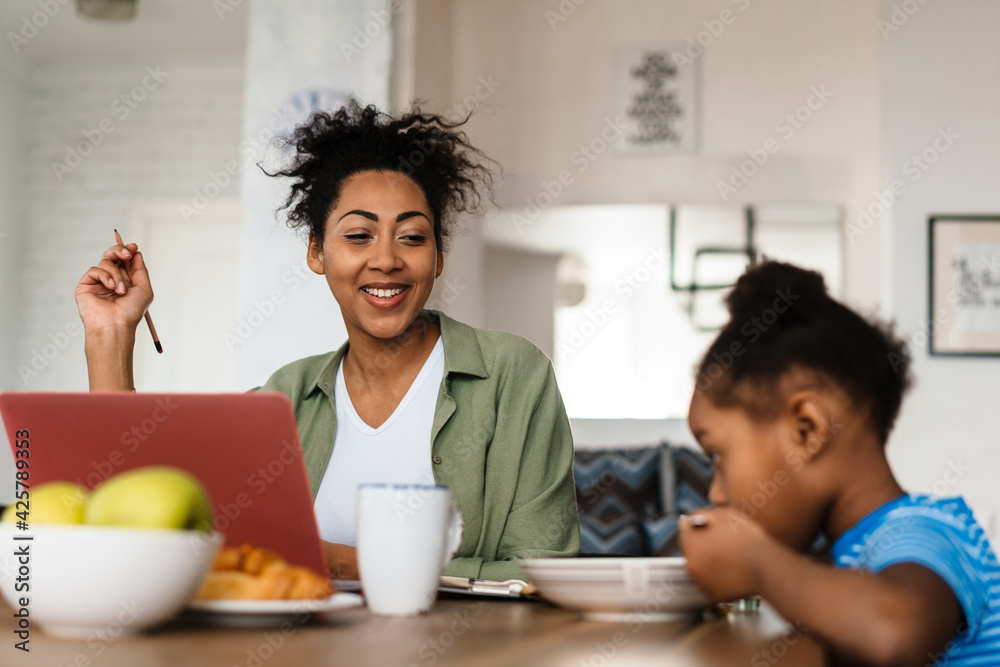 Black smiling woman working while having breakfast with her daughter Black smiling woman working while having breakfast with her daughter