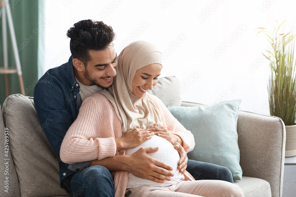 Happy islamic couple expecting baby hugging on couch at home