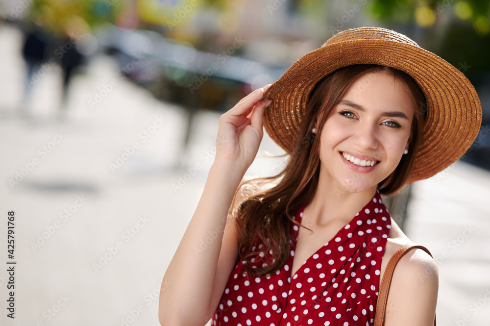 Young happy smiling woman (female) in red polka dot dress and straw hat walks in the summer city