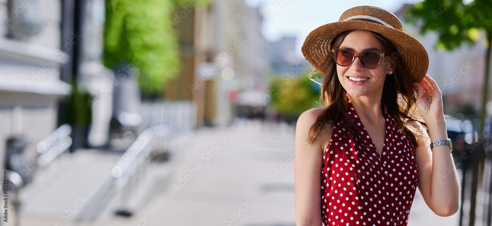 Young smiling girl (female) in sunglasses straw hat and red dress with polka dots walks in the summer around the city