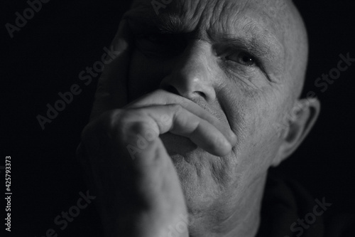 Close up portrait of a mature make with hand over his moth looking puzzled and frowning. Black and white photography with a head shot against a black background