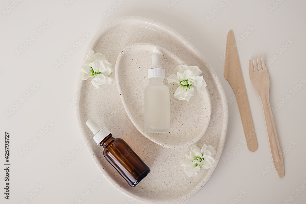Two glass bottles for cosmetic, natural medicine , essential oil inside avocado shaped ceramic plates decorated with flowers and a cutlery set on a white background. Top view, beauty product flat lay. Two glass bottles for cosmetic, natural medicine , essential oil inside avocado shaped ceramic plates decorated with flowers and a cutlery set on a white background. Top view, beauty product flat lay.