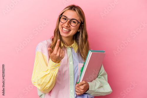 Young caucasian blonde student woman holding books isolated pointing with finger at you as if inviting come closer.