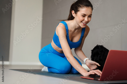 Smiling woman playing with her dog and using laptop during yoga practice