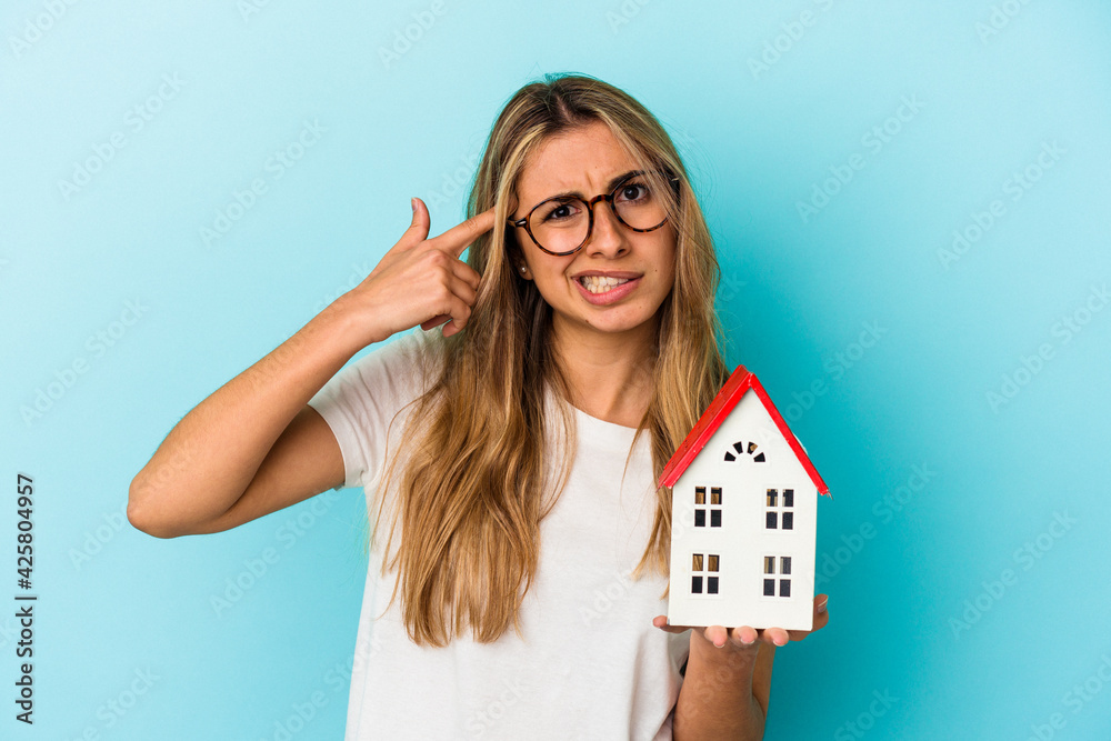 Young caucasian woman holding a house model isolated on blue background showing a disappointment gesture with forefinger. Young caucasian woman holding a house model isolated on blue background showing a disappointment gesture with forefinger.