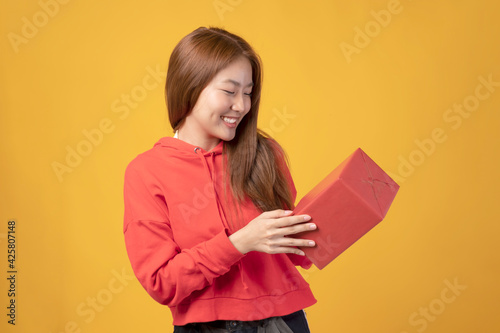 Portrait of casual young happy smiling Asian woman hold gift box. Beautiful young Asian girl holding red gift box over yellow background. Cute Asian girl with red jacket holding gift box.