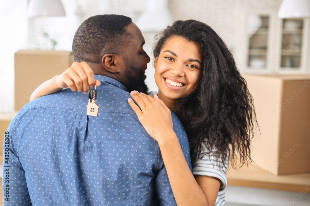 Obraz premium Close photo of delighted couple who bought new flat, black man standing with his back and hugging his happy wife with the keys to their new apartment, buying a property, a new stage in a relationship