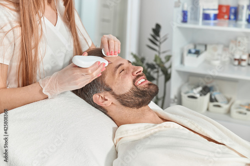 Bearded man smiling in professional beauty spa salon during ultrasonic facial cleansing procedure
