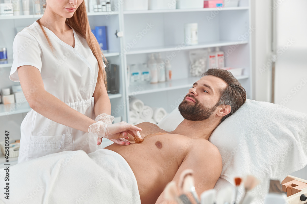 Fototapeta premium Woman beautician put depilatory wax to chest of young man for hair removal. Depilation with wax