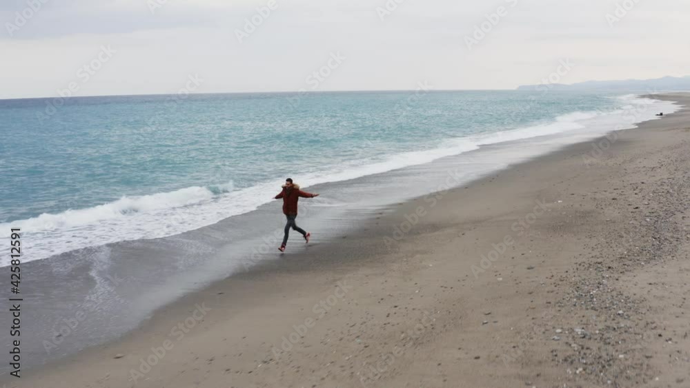 Giovane ragazzo corre con il cappotto sulla spiaggia con mare in tempesta Giovane ragazzo corre con il cappotto sulla spiaggia con mare in tempesta