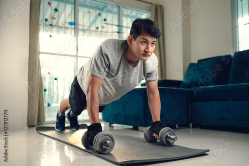Young man doing push up with dumbbell and exercises on yoga mat in living room at home. Fitness, workout and traning at home concept.