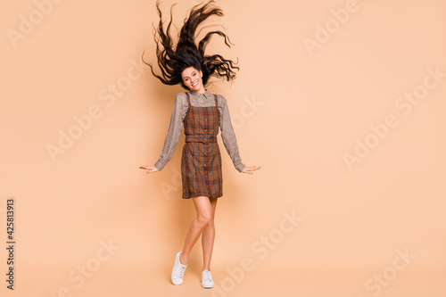 Full length body size view of pretty cheery girl wearing checked uniform having fun throwing wavy hair isolated on beige pastel color background