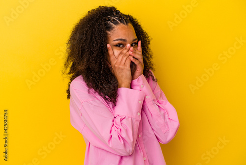 Young african american woman isolated on yellow background blink through fingers frightened and nervous.