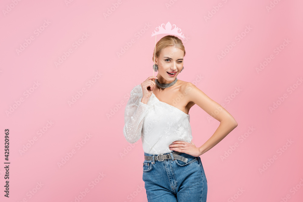 blonde woman holding paper crown on stick and standing with hand on hip isolated on pink blonde woman holding paper crown on stick and standing with hand on hip isolated on pink