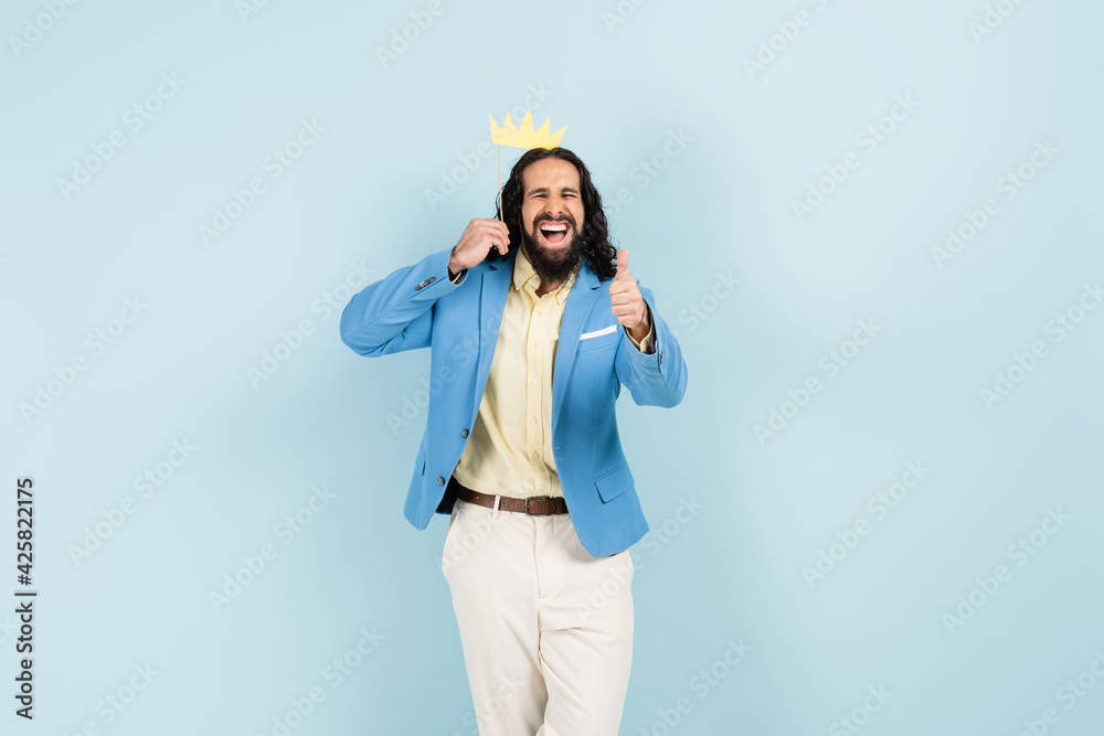 happy hispanic man in jacket holding paper crown on stick and showing thumb up isolated on blue happy hispanic man in jacket holding paper crown on stick and showing thumb up isolated on blue