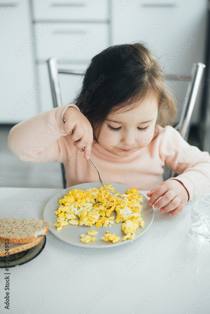 Baby girl in the afternoon in the white light kitchen in a pink sweater eating an omelet Baby girl in the afternoon in the white light kitchen in a pink sweater eating an omelet