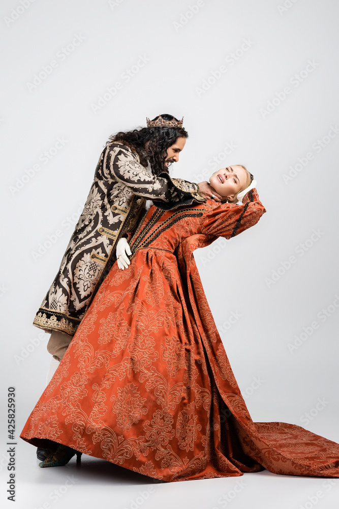 full length of cruel hispanic king in medieval clothing choking blonde queen in golden crown on white