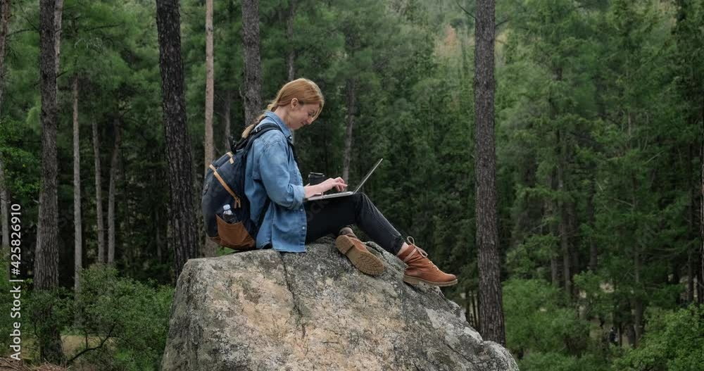 Young freelancer woman with backpack working on the laptop in the forest sitting on huge rock. Young freelancer woman with backpack working on the laptop in the forest sitting on huge rock.