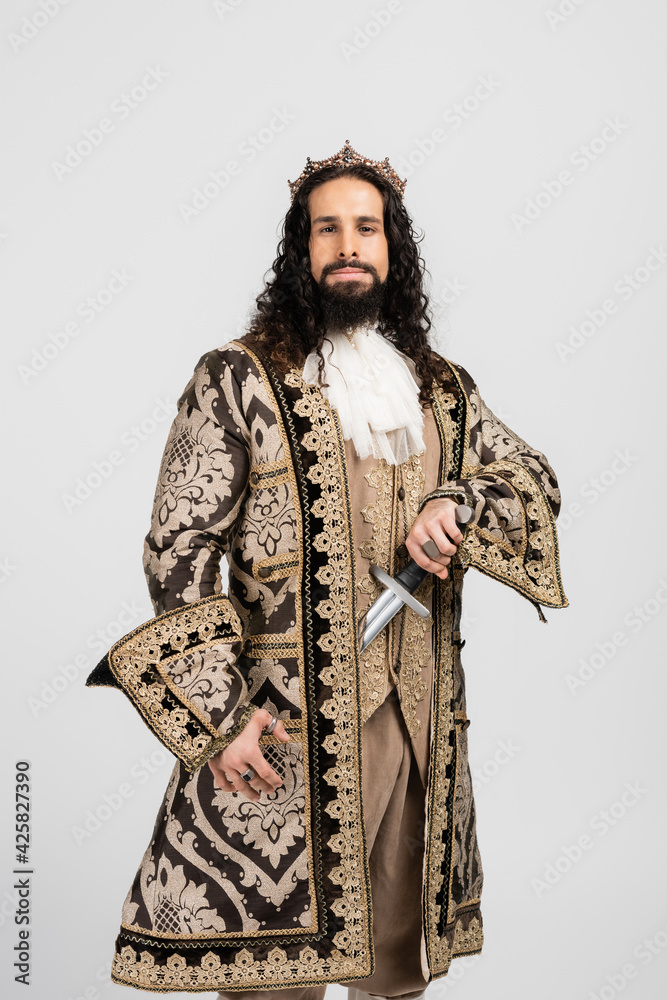 hispanic king in golden crown and medieval clothing holding sword while looking at camera isolated on white