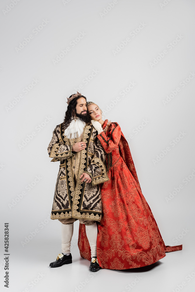full length of blonde queen in royal crown leaning on hispanic king in medieval clothing on white