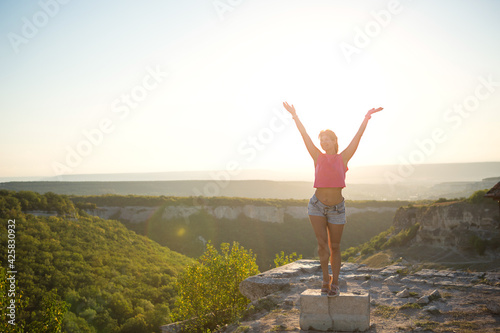 Female tourist with her hands raised looks at panoramic view on the top of the mountain and rejoices, enjoys it freedom and adventure. Trekking, travel, active ecotourism, healthy lifestyle, hiking