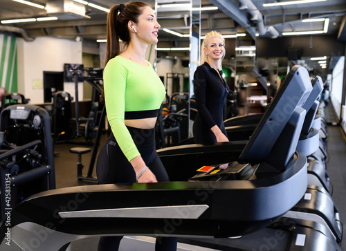 Beautiful smiling athletic woman doing fitness cardio exercises in the gym, running on a treadmill. Two sporty women working out together