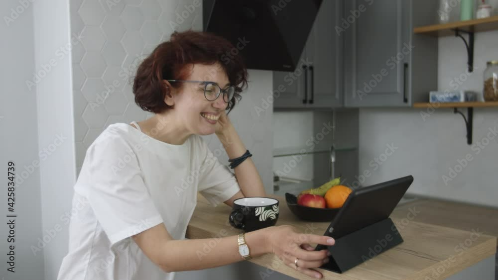 Smiling young woman sitting in kitchen, drinking tea and talking video call, having online conference, conversation with friend via digital tablet while being lockdown quarantine, pandemic concept Smiling young woman sitting in kitchen, drinking tea and talking video call, having online conference, conversation with friend via digital tablet while being lockdown quarantine, pandemic concept