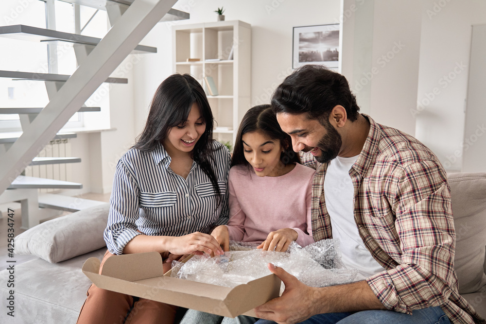 Excited indian family with child daughter unpack parcel at home. Happy parents and teen kid daughter open postal box receive present gift in online shopping delivery package sitting on sofa together. Excited indian family with child daughter unpack parcel at home. Happy parents and teen kid daughter open postal box receive present gift in online shopping delivery package sitting on sofa together.