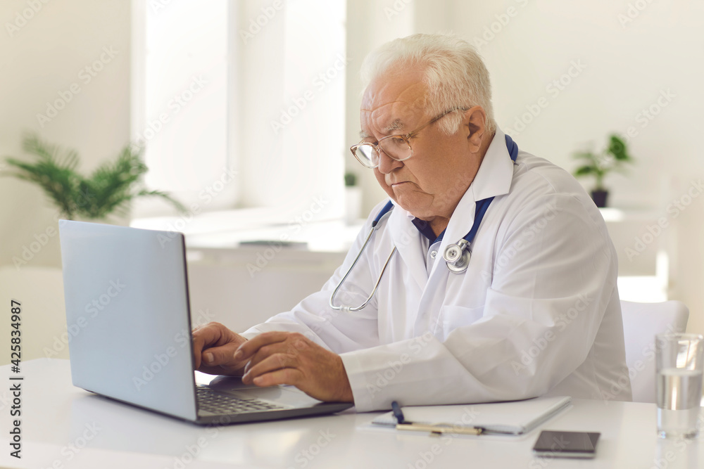 White-haired senior doctor typing on laptop keyboard sitting at desk in hospital office White-haired senior doctor typing on laptop keyboard sitting at desk in hospital office