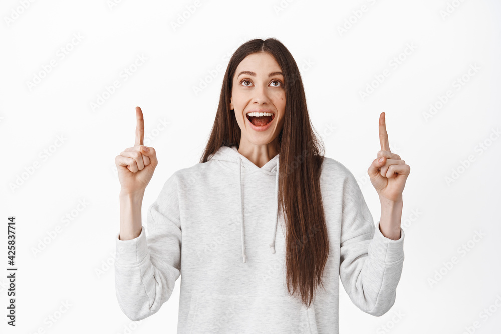 Fascinated young woman checking out promotional text, pointing and looking amazed at banner logo, super cool deal offer, standing in hoodie against white background Fascinated young woman checking out promotional text, pointing and looking amazed at banner logo, super cool deal offer, standing in hoodie against white background