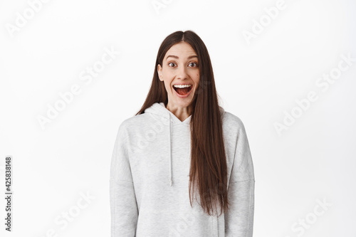 Surprised and thrilled girl smiling, standing in awe with opened mouth and looking at camera fascinated, breathaking and exciting event, posing against white background photo