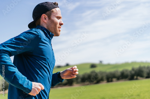 A young white european and athletic man is running in the countryside on a sunny day. The guy is wearing blue clothes and a cap.