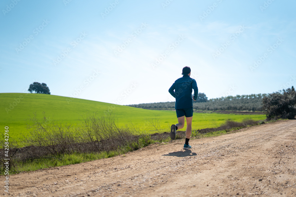 A young white european and athletic man is running in the countryside on a sunny day. The guy is wearing blue clothes and a cap.