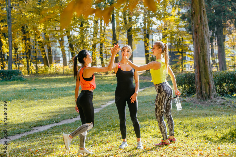 Fototapeta premium Full height of group of three female athletes give each other high five after a good training session in fall park. Attractive sporty caucasian woman with two young fitness friends smiling outdoor.