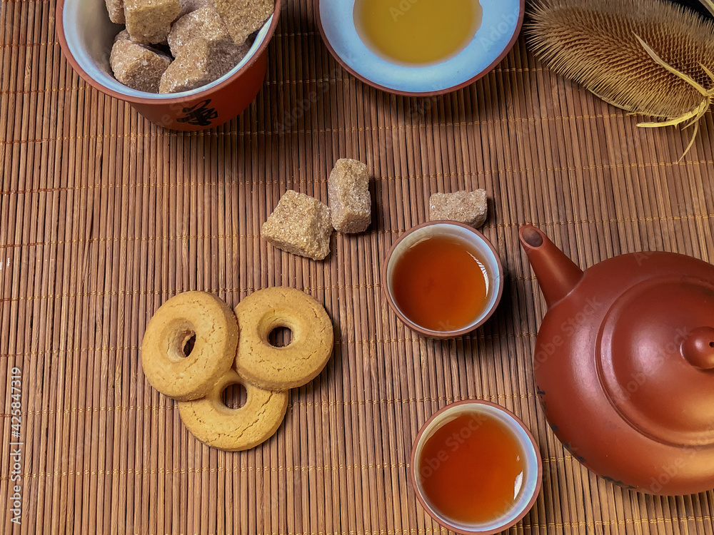 Brown chinese tea set of a teapots and two cups, cookies and brown sugaron on bamboo table mat . Close- up photo, view from the top. Brown chinese tea set of a teapots and two cups, cookies and brown sugaron on bamboo table mat . Close- up photo, view from the top.