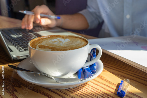 A white mug of cappuccino with a nice blurry hand typing on a laptop in the background