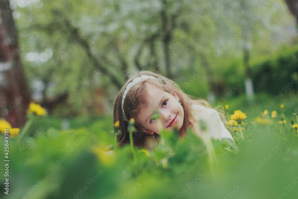 A beautiful girl in white hiding in grass in spring garden. Portrait of child among green grass and yellow dandelions. Young lady sitting in sunny blooming park and smilling. A beautiful girl in white hiding in grass in spring garden. Portrait of child among green grass and yellow dandelions. Young lady sitting in sunny blooming park and smilling.
