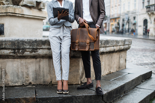 Close up of afro american woman with clipboard and young man with briefcase sitting together outdoors. Two business colleagues solving urgent working issues on street.