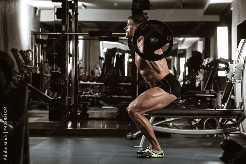 Fototapeta premium Fitness in gym, sport and healthy lifestyle concept. Handsome athletic man with naked torso making exercises. Bodybuilder male model training muscles making squats with barbell. Side view