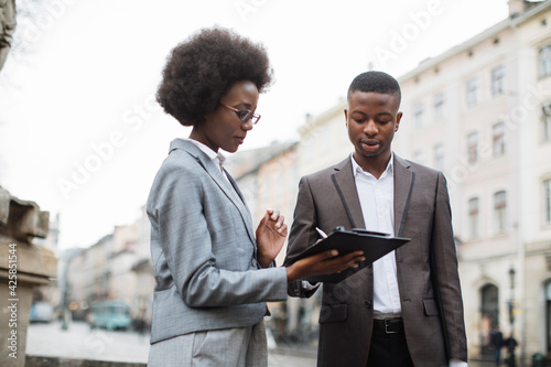 Portrait of two busy african people looking on clipboard while standing on city street. Female assistant helping her male employer to make schedule for working week. © sofiko14