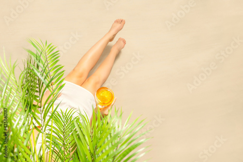 Alone woman sitting under palm tree branches with glass of water with piece orange. Female relaxation on the sand of the beach at summer vacation. Top view